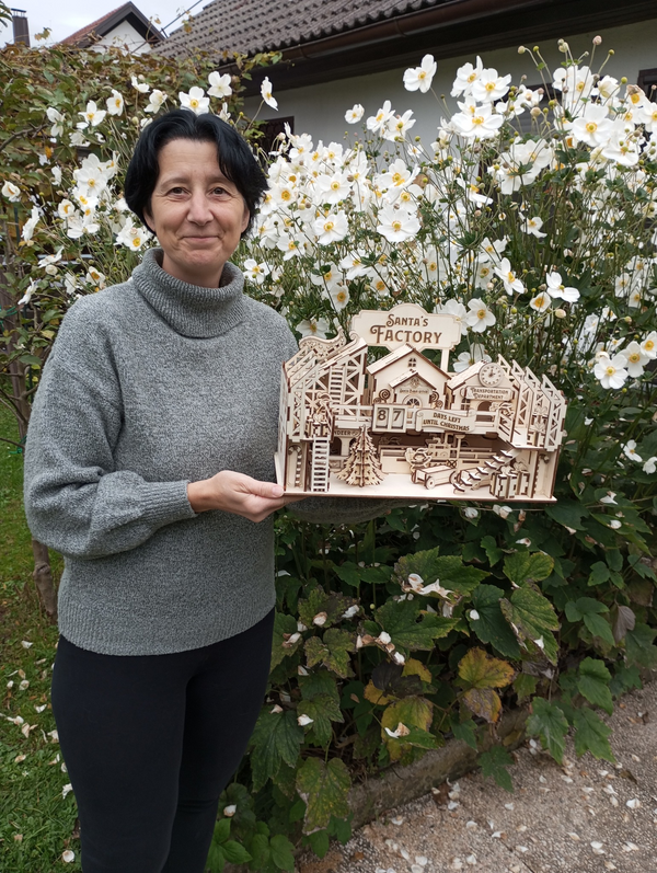 Klara Kolenc Babic holding a wooden model of a factory made by Sculpfun ci mini laser engraver  in front of a house with white flowers.
